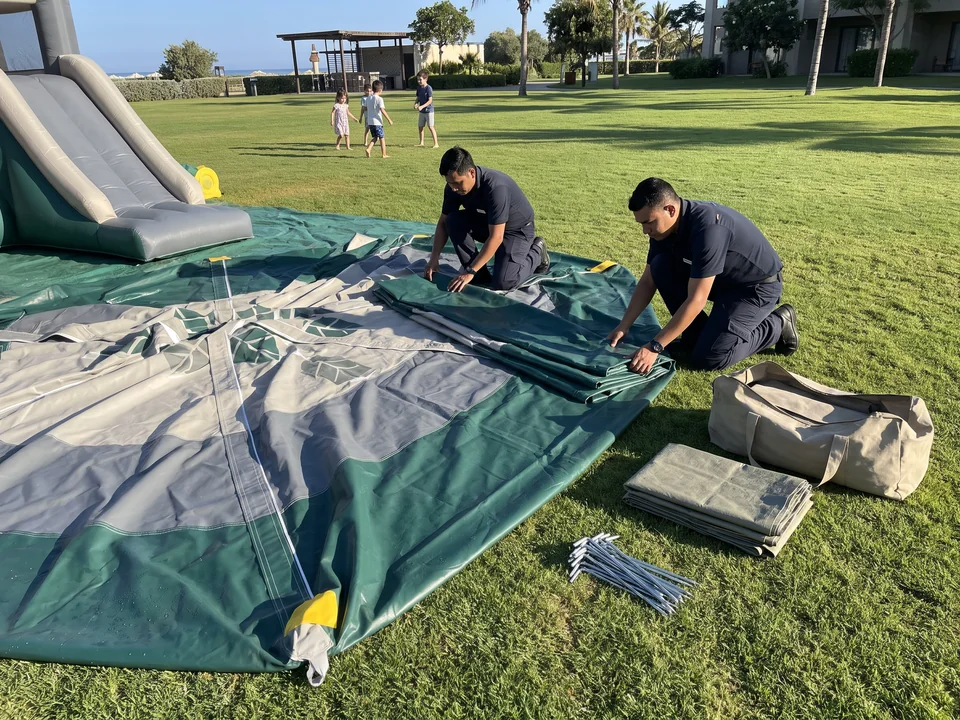 Two staff packing an inflatable on a resort lawn, its storage bag and ground stakes beside them