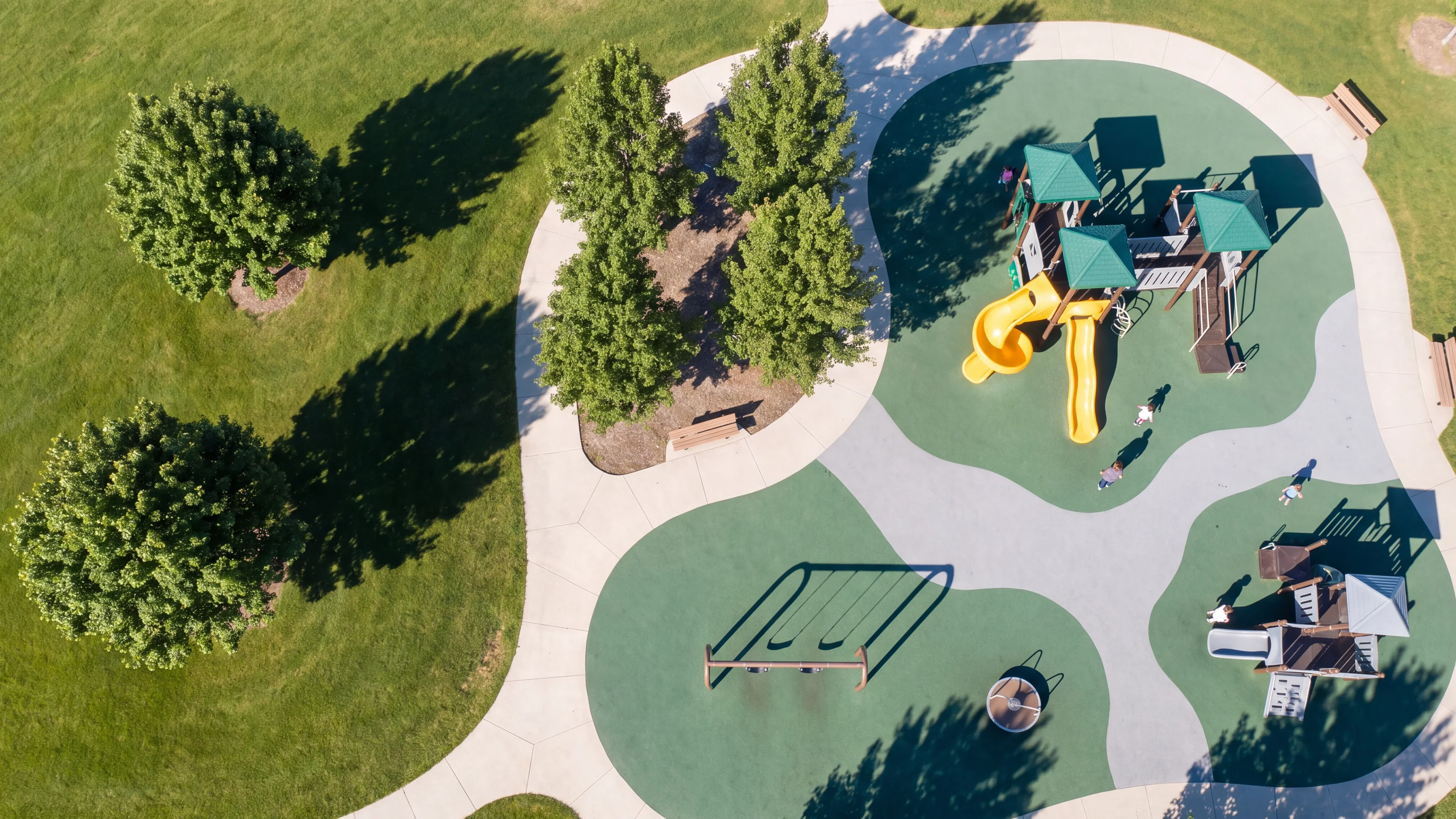 Straight-down aerial of a park playground with three structures, a swing set, a spinner and evergreen and grey rubber surfacing