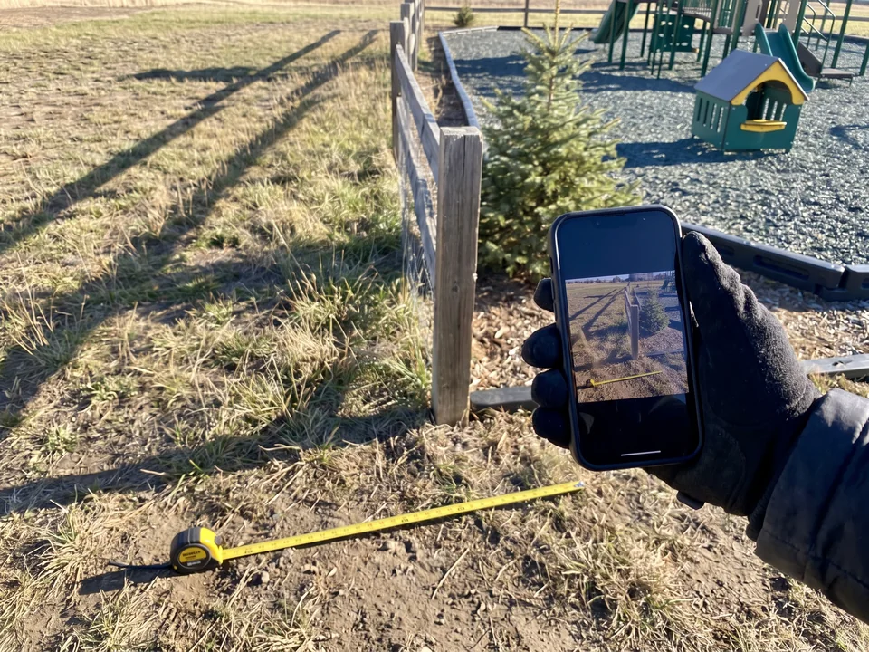 A hand photographing an empty site corner with a phone, a tape measure stretched behind