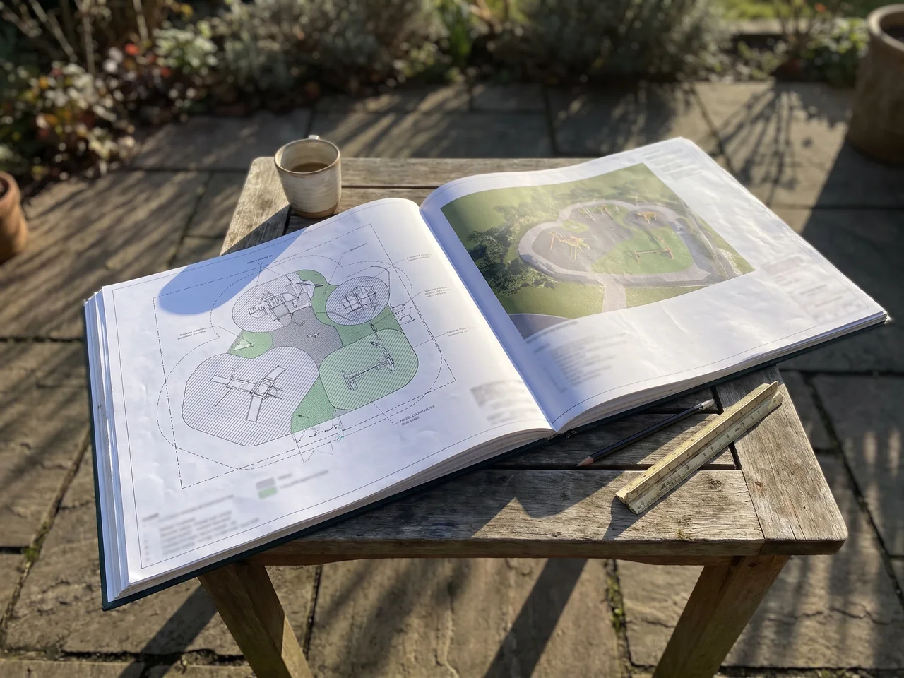 A bound plan set open at the playground sheet beside a printed aerial render