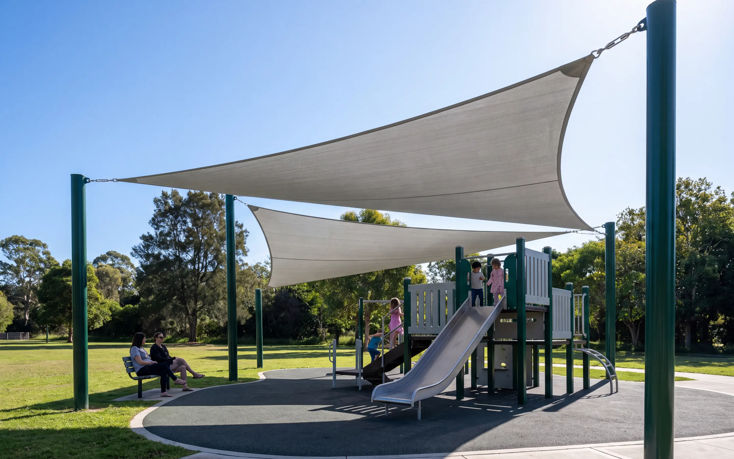 Triangular shade sail on three evergreen posts casting a crisp shadow over a multi-deck play structure in morning light