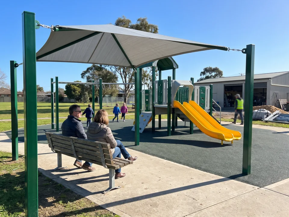 A small bench shade on two evergreen posts over a park bench, two parents sitting in its shadow