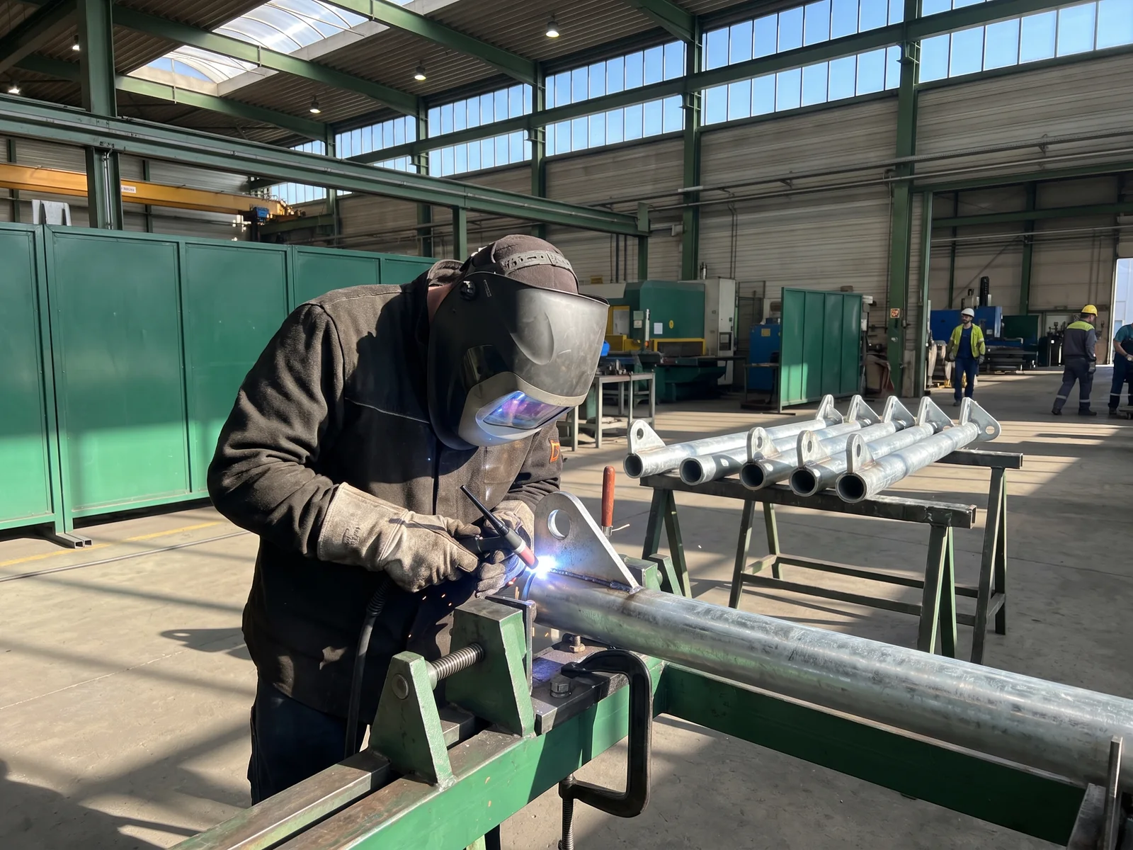 A welder welding the eye plate onto the top of a galvanized shade post on a jig in our steel workshop