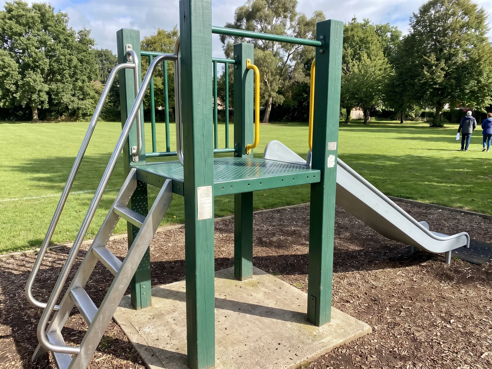 A freestanding slide unit in a park corner: ladder, punched deck on evergreen posts and a grey straight slide